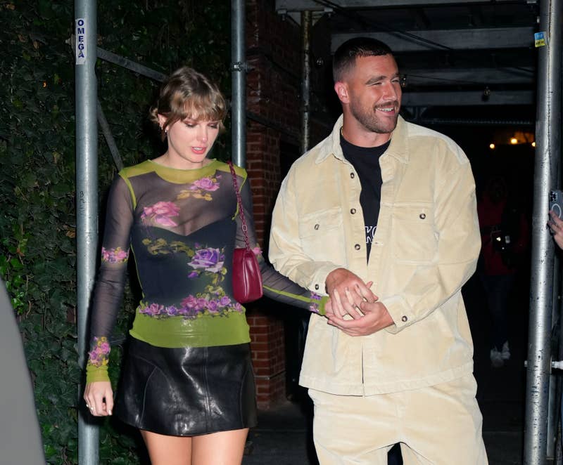 Taylor Swift in a floral top and leather skirt walks alongside a man in a light jacket and dark pants