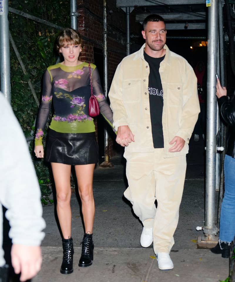 Taylor Swift in a sheer floral top and leather skirt with boots alongside Joe Alwyn in a beige jacket and pants, walking hand in hand