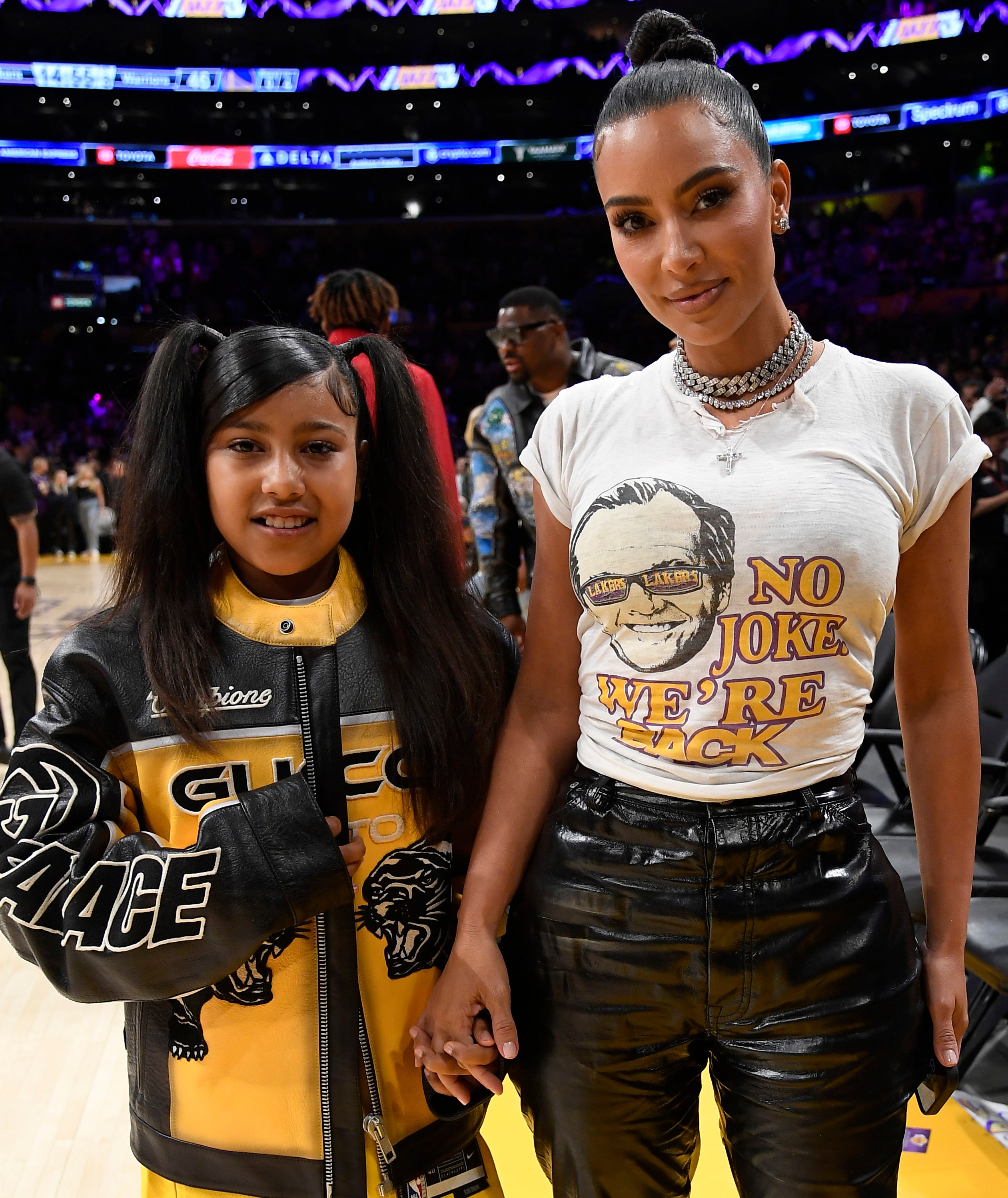 Kim, in graphic tee and shiny pants, holding hands with North in a sports jacket, both standing on a basketball court