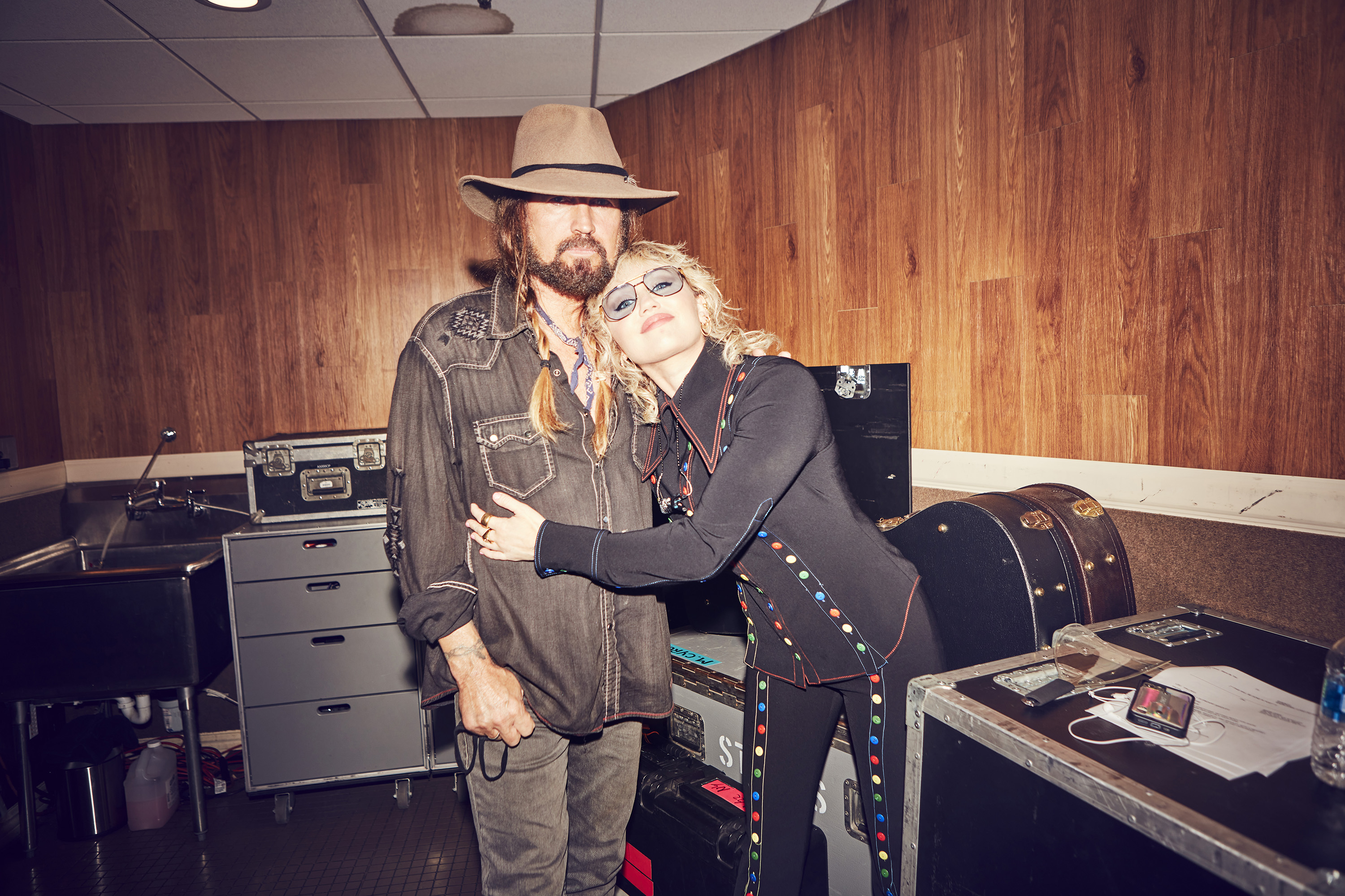 Miley, in a black outfit with ribbon details, hugs Billy Ray, in a hat and denim jacket, backstage
