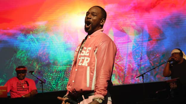 Performer in a pink jacket and chain singing on stage with band members and colorful background
