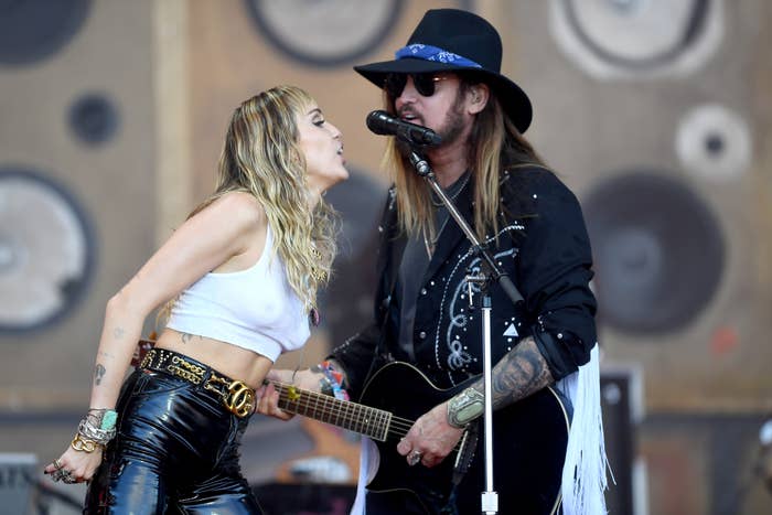 Miley Cyrus and Billy Ray Cyrus performing onstage, Miley wearing a crop top and Billy Ray in a jacket and hat