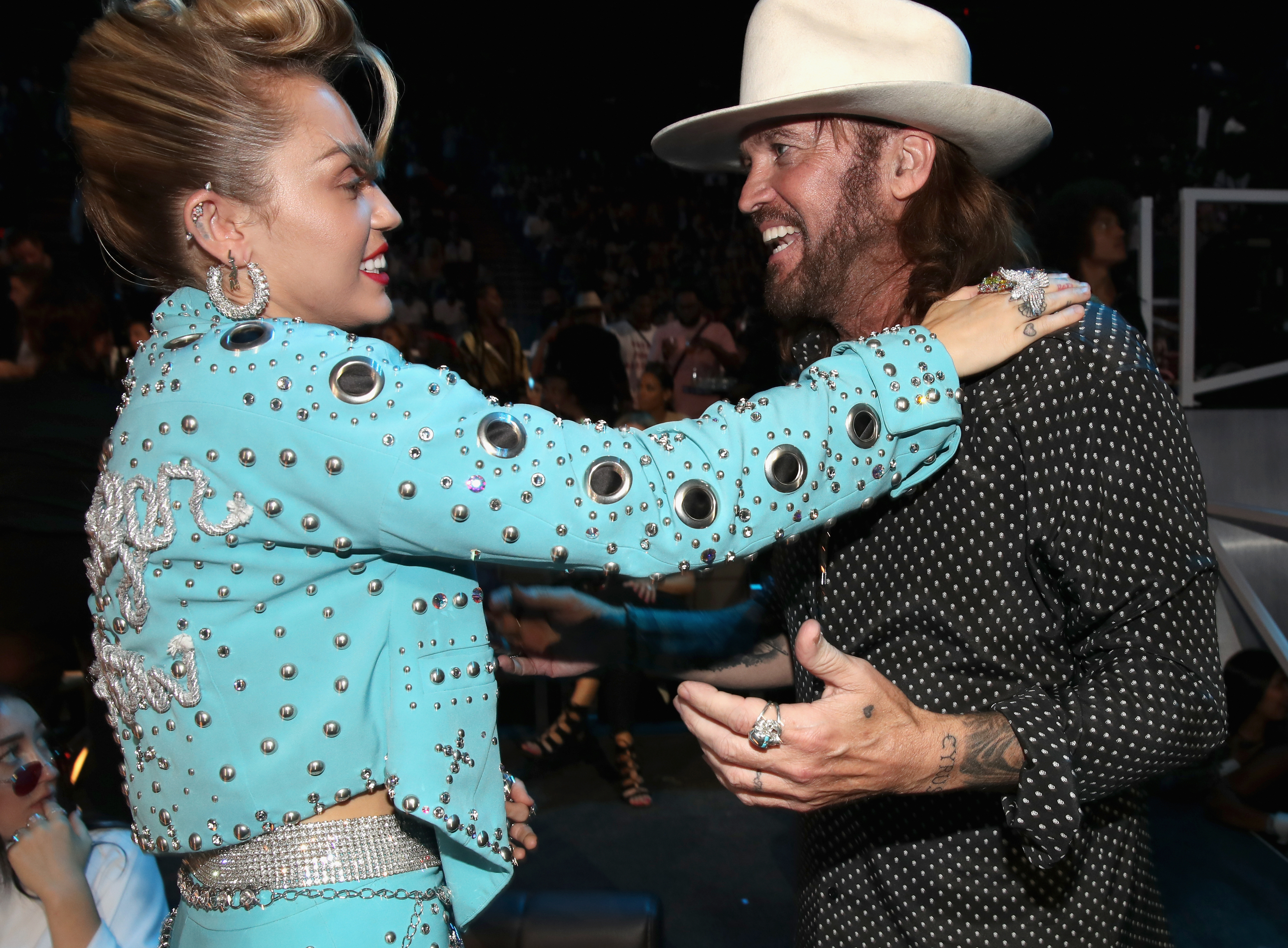 Miley Cyrus in embellished blue jacket hugging Billy Ray Cyrus in a hat and polka-dot shirt at event