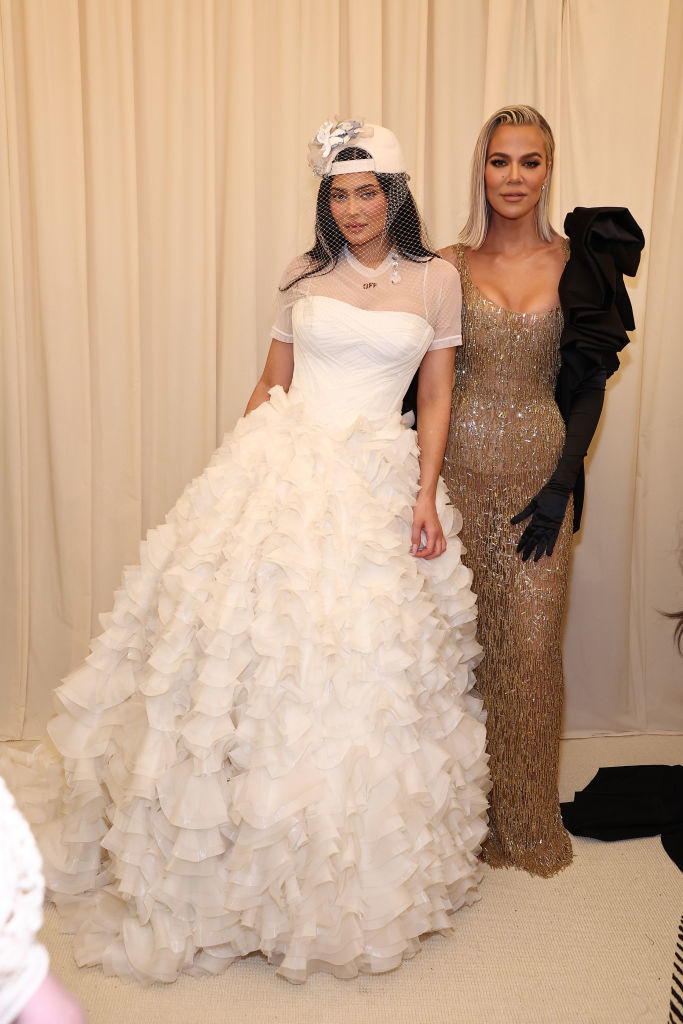 the two sisters in elegant attire, Kylie in a layered gown and headpiece, Khloe in a sequined, form-fitting dress with a glove