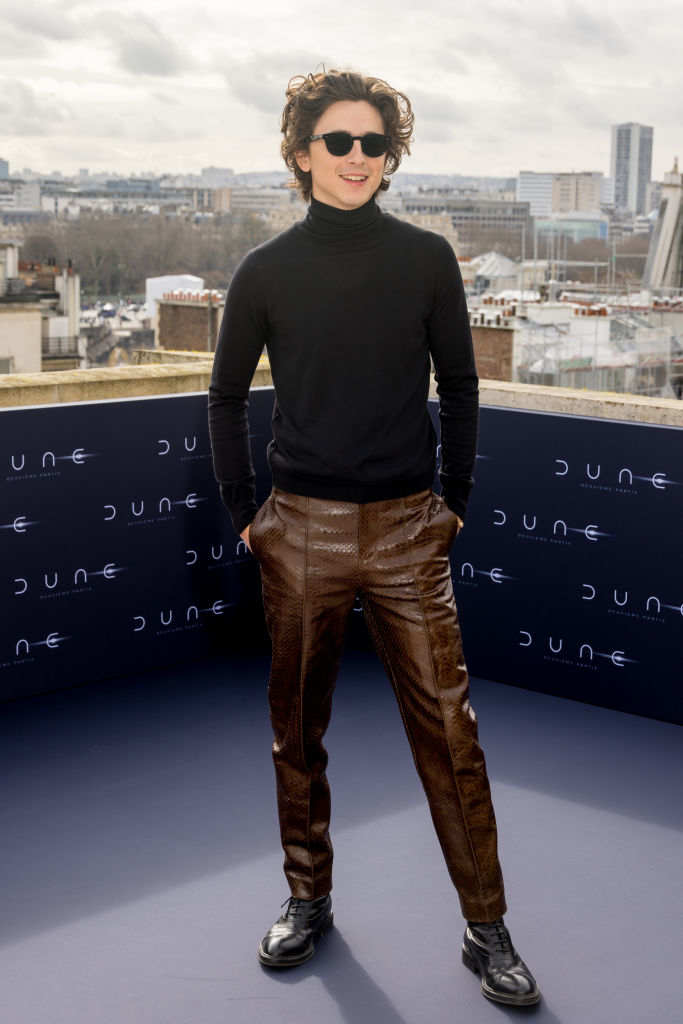 Timothée stands on a terrace smiling, sporting a black turtleneck and brown textured pants and wearing sunglasses