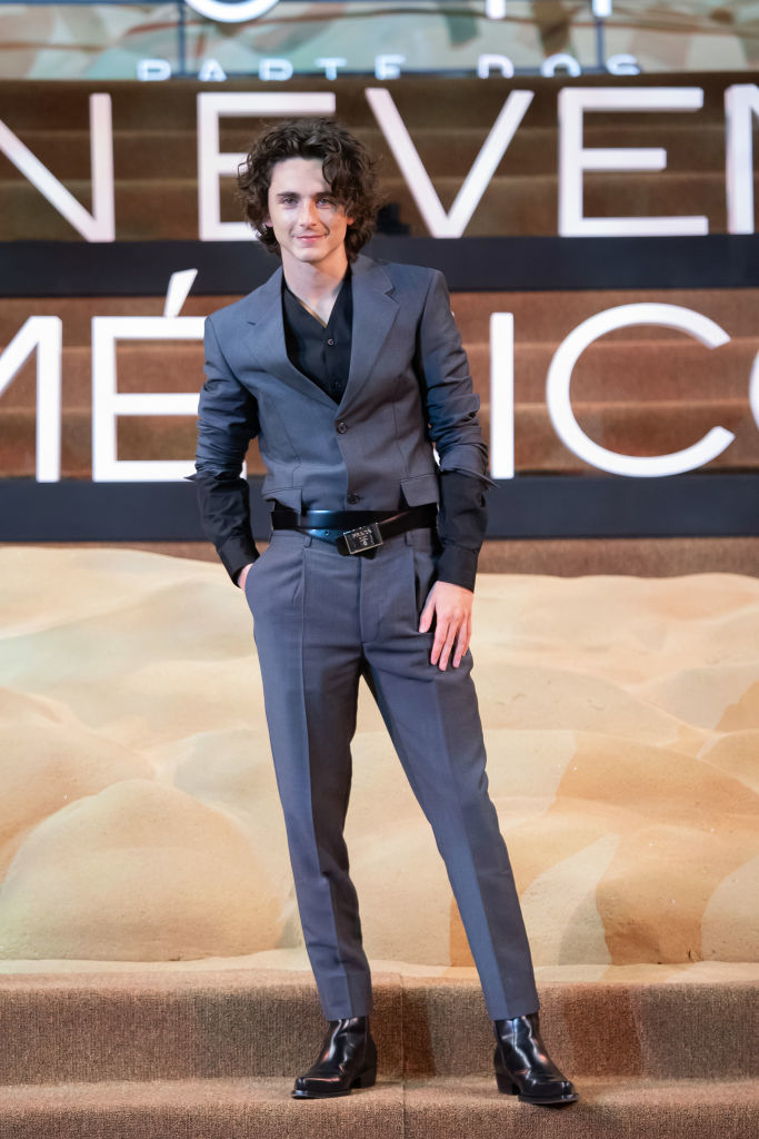 Timothée standing on a stage, wearing a tailored suit and a slight smile, hand in pockets