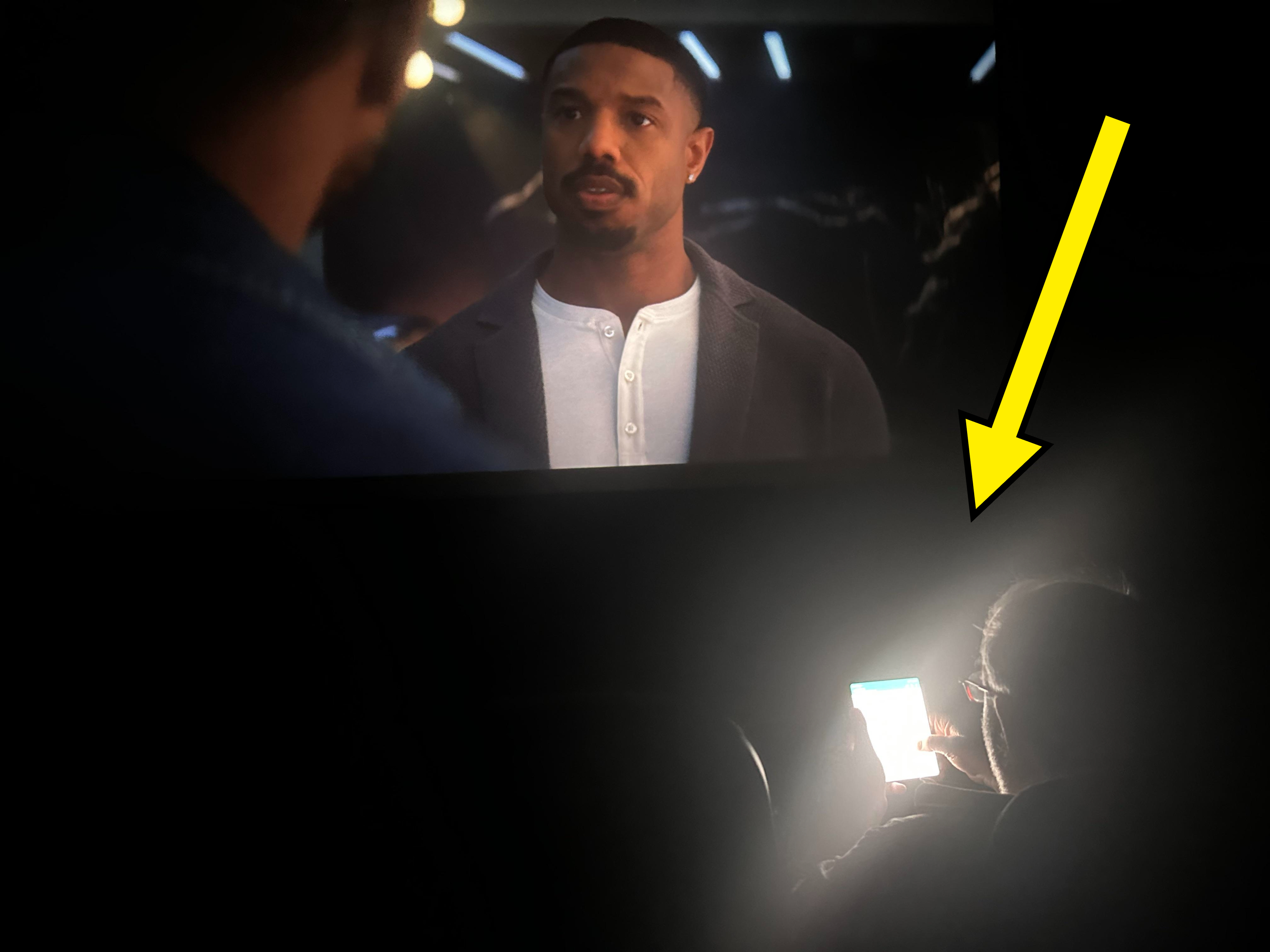 Person holding a phone with bright screen in a dark cinema, distracting from a movie scene with a man speaking