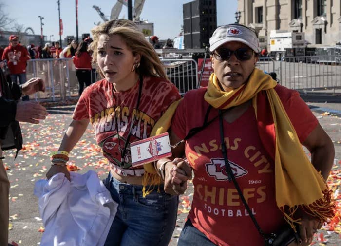 Two fans in Kansas City Chiefs attire walk through a strewn confetti area, one carrying memorabilia