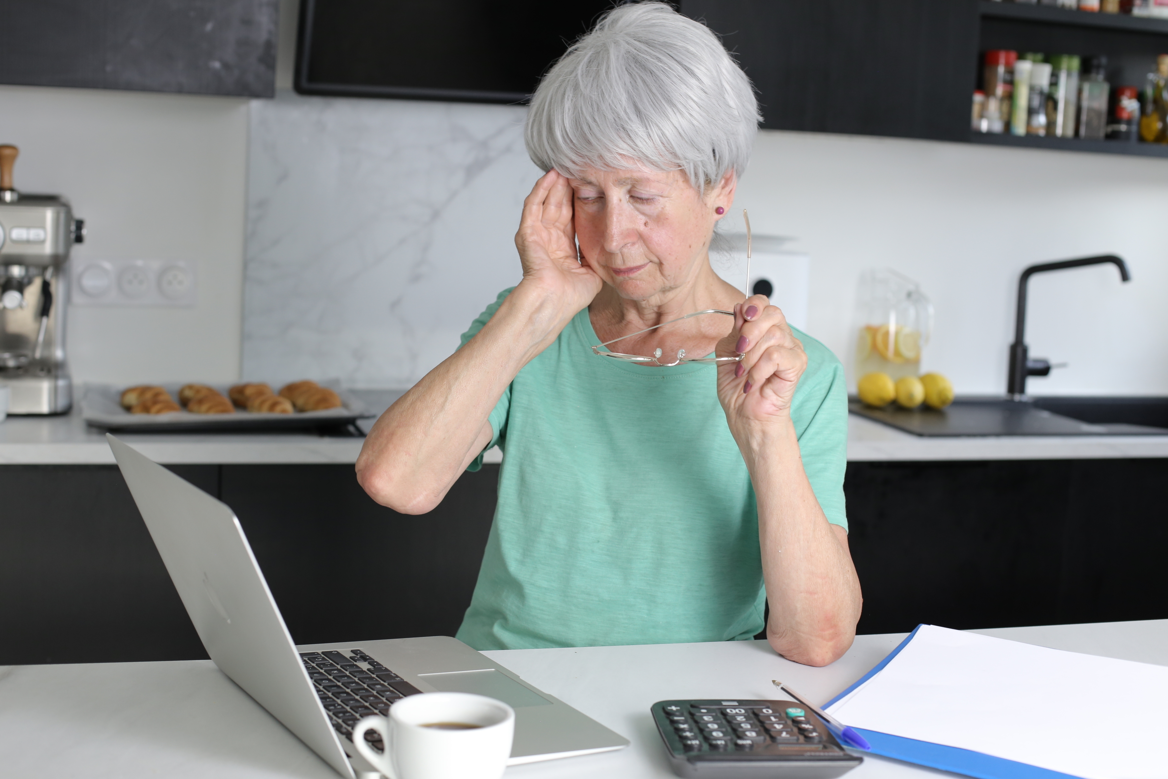 Older woman with glasses appears frustrated or tired while working on a laptop in a kitchen setting