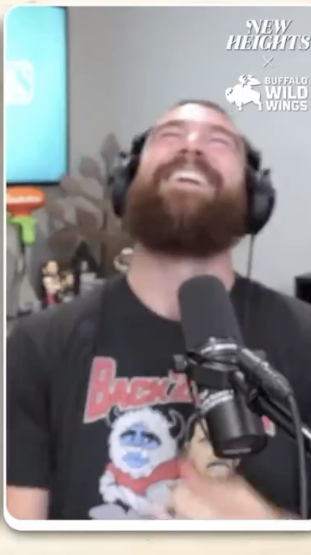 Travis, in a graphic T-shirt and wearing headphones, laughing in front of a microphone in a room with toys in the background