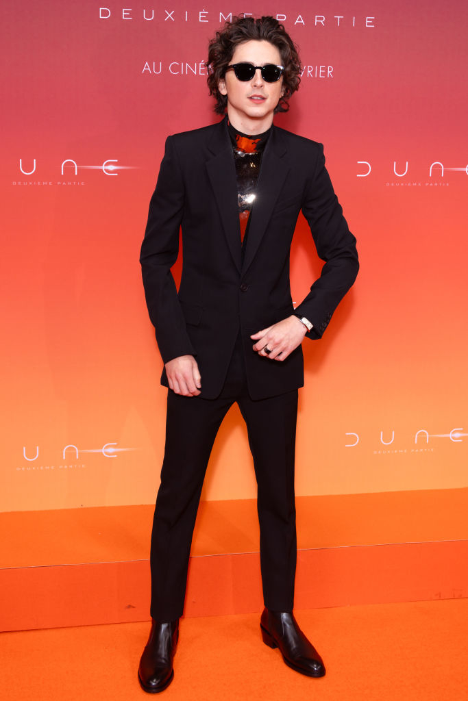 Timothée in a black suit with hands in pockets standing against an orange backdrop