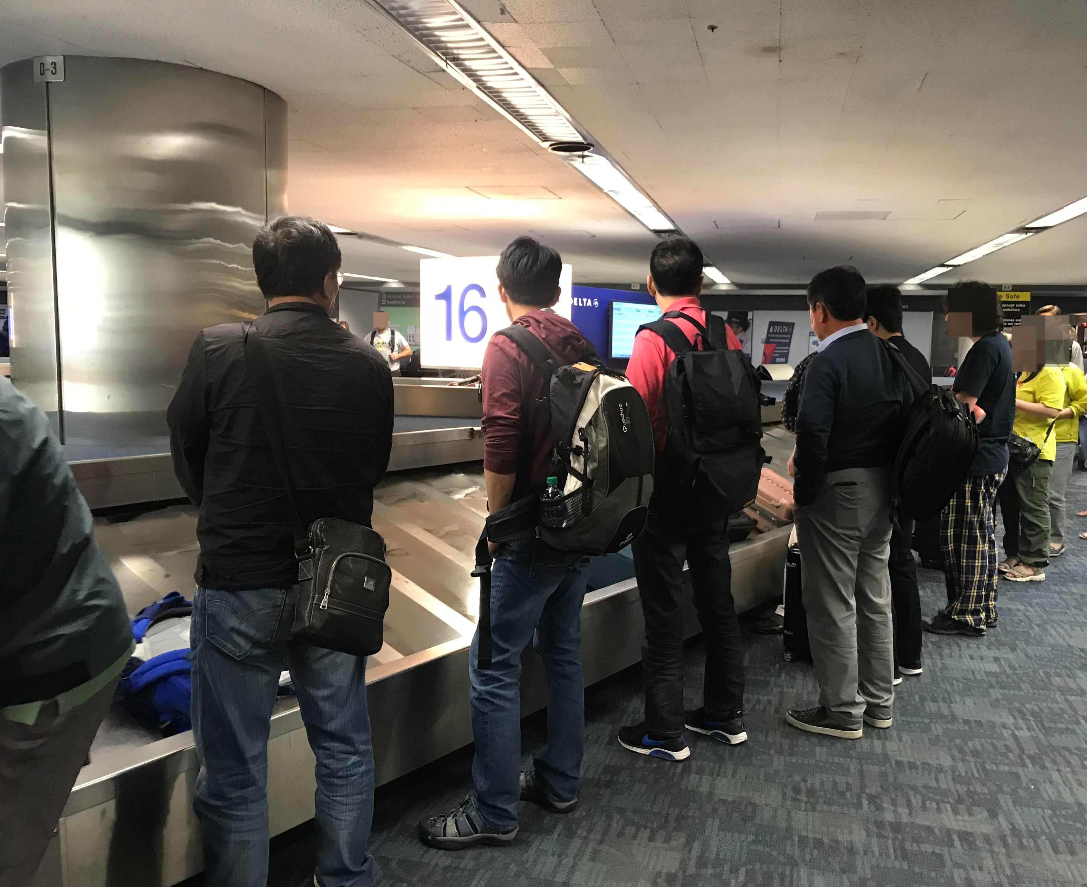 Passengers standing by a baggage carousel, waiting for luggage; some with backpacks