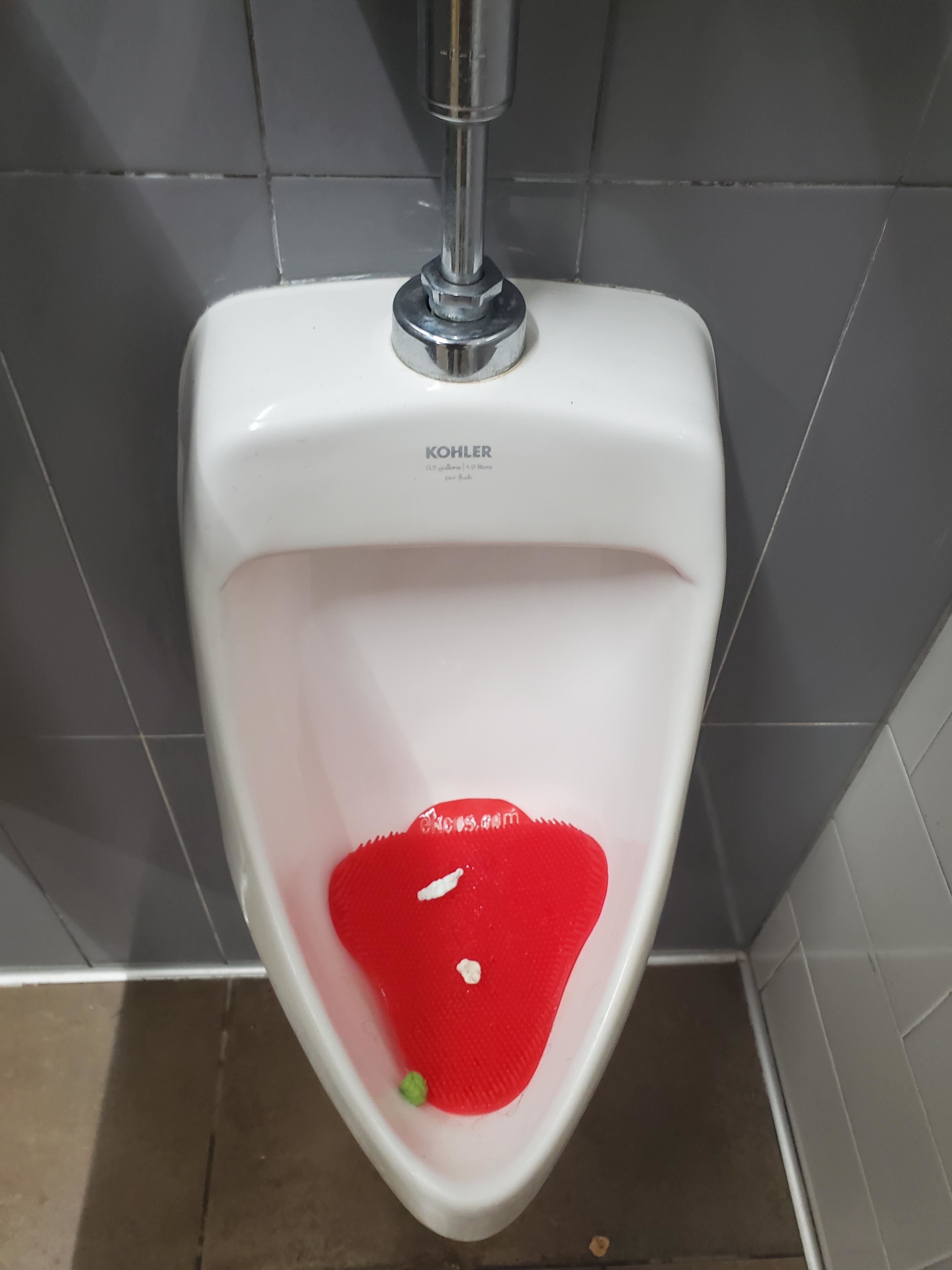 Urinal with a red deodorizer block inside that people threw gum into, against a gray tiled wall