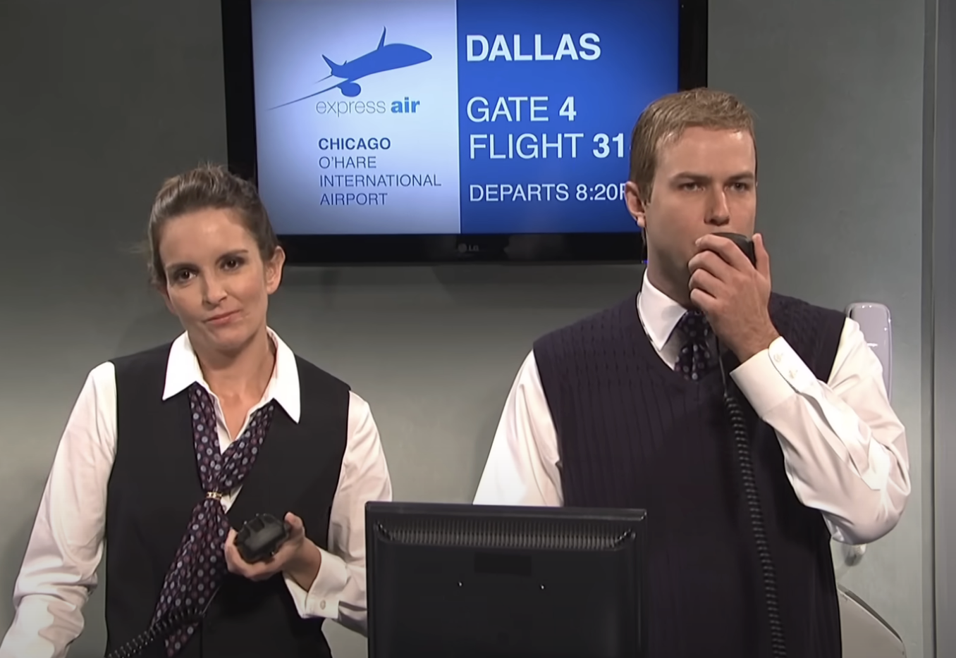 Two actors in airport staff uniforms stand at a gate podium, one holding a microphone, in a comedic sketch
