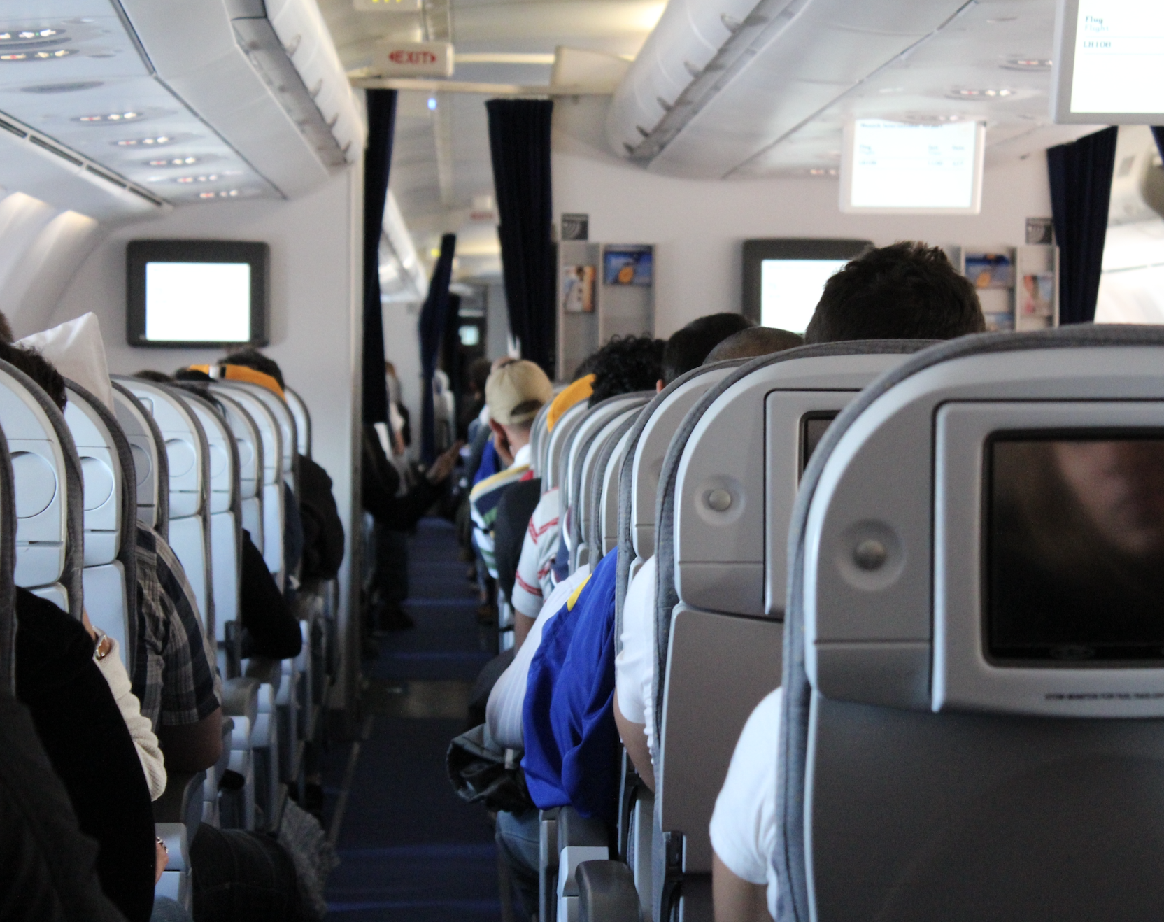 Passengers seated in an airplane cabin, some watching overhead screens