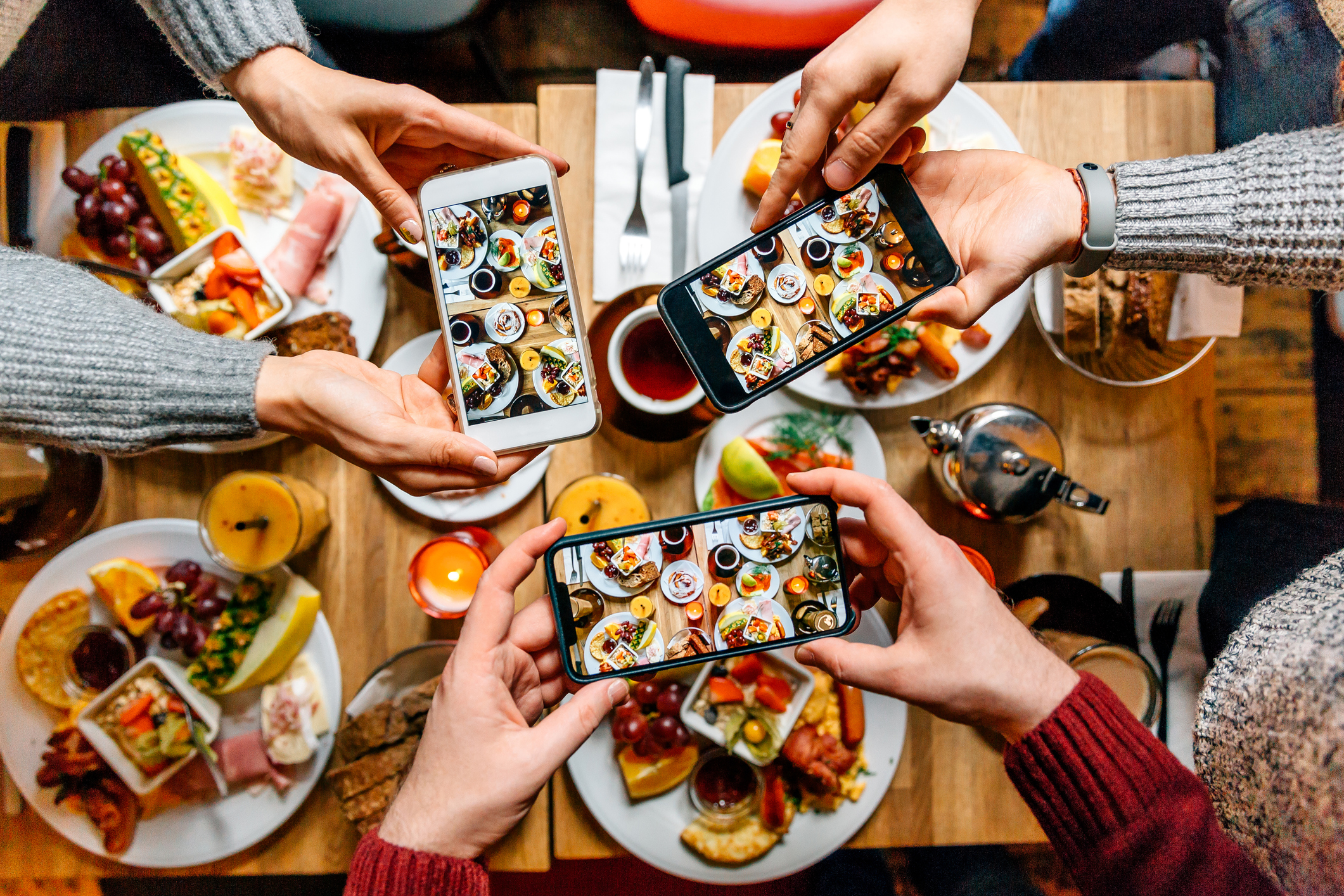 Four hands holding smartphones above a table filled with various dishes, taking photos of the food