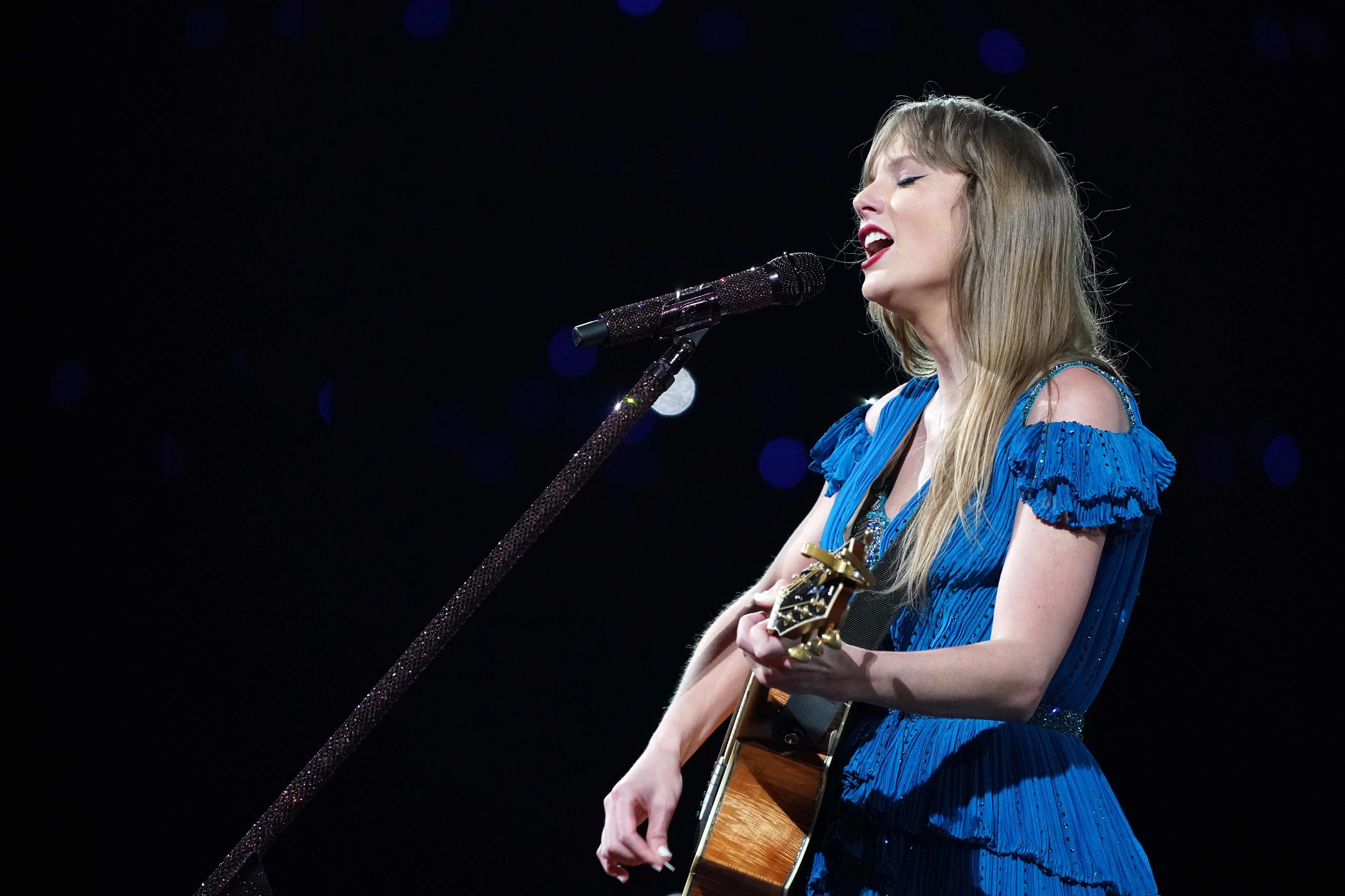 Taylor in a blue fringe dress playing guitar and singing into a microphone onstage