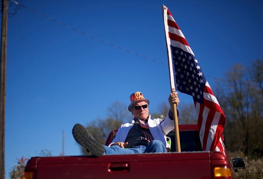 Person in a decorated hat and sunglasses sits in pickup bed with an American flag