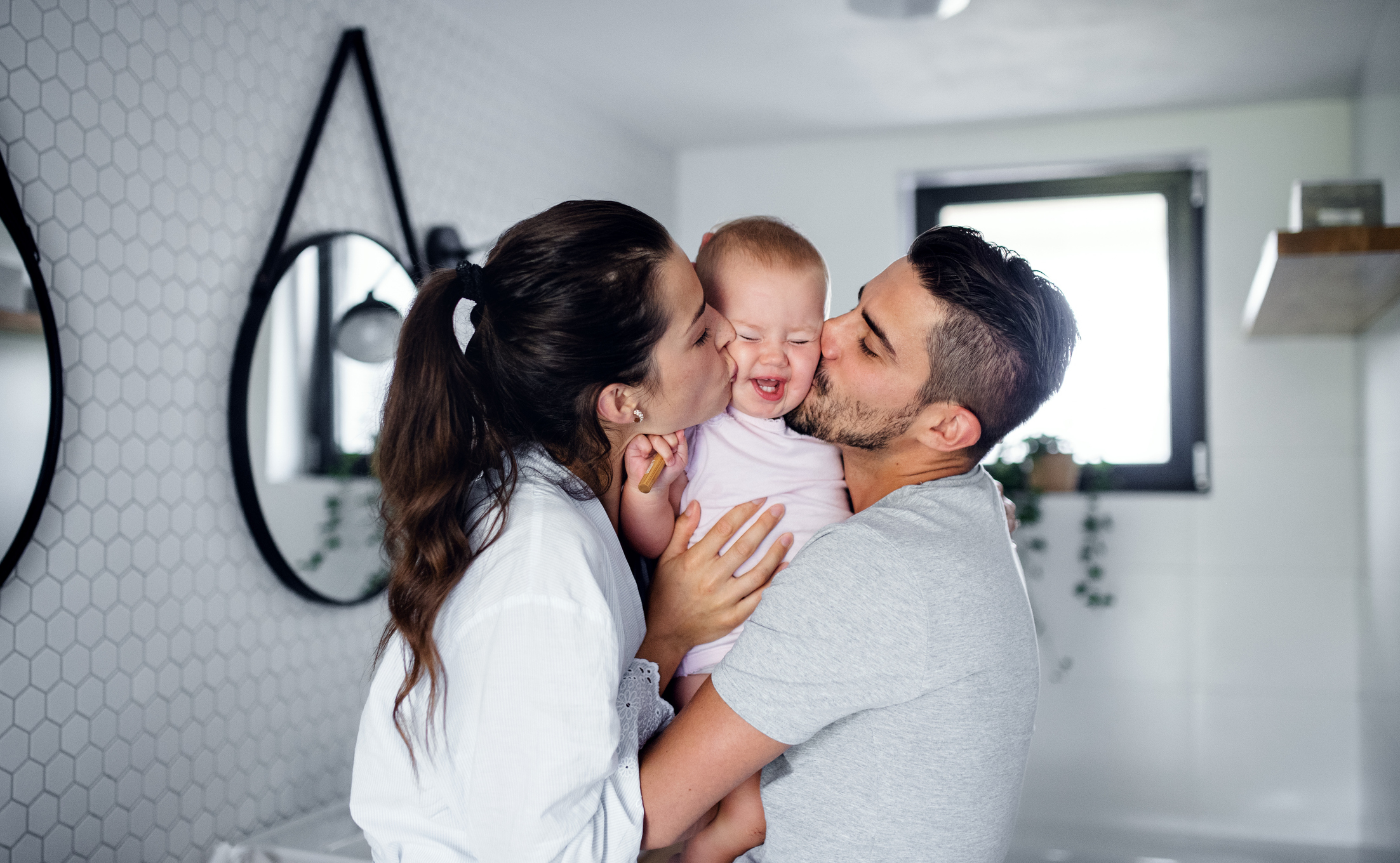 Two parents lovingly kiss a toddler&#x27;s cheeks from either side in a home bathroom