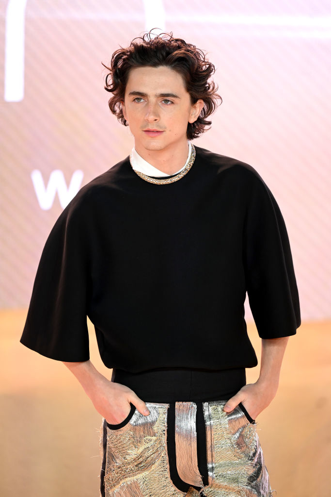 Timothée standing with hands on hips wearing a black top, chunky necklace, and patterned trousers