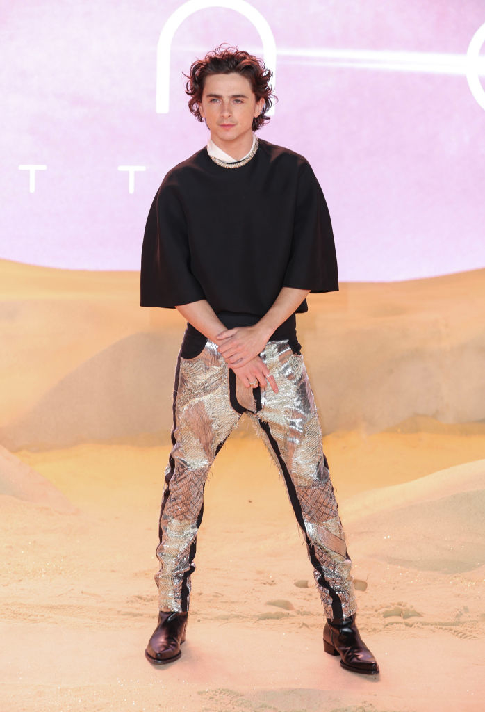 Timothée in a black top and shiny metallic pants posing on an event backdrop