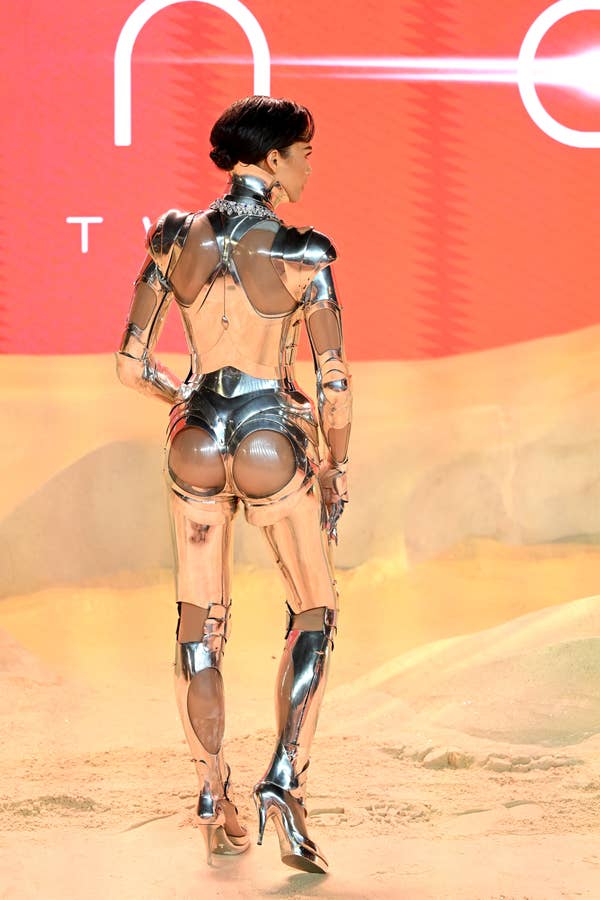 Zendaya in a metallic structured outfit with reflective surfaces, posing on a sandy stage with branding in the background