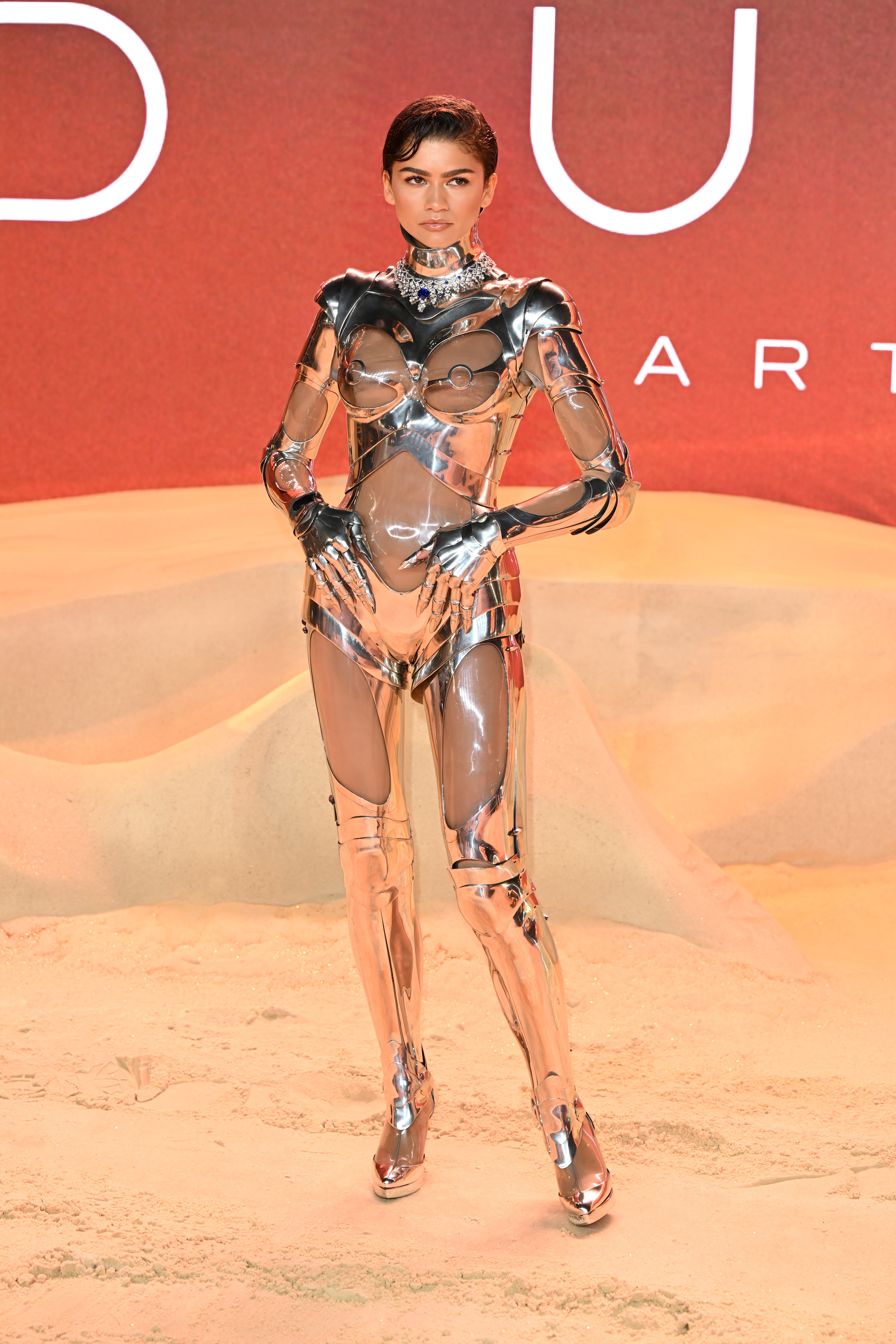 Zendaya in a futuristic metallic bodysuit stands in front of a branded backdrop
