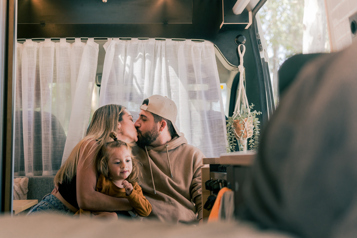 Two adults kiss while embracing a young child in a cozy RV interior