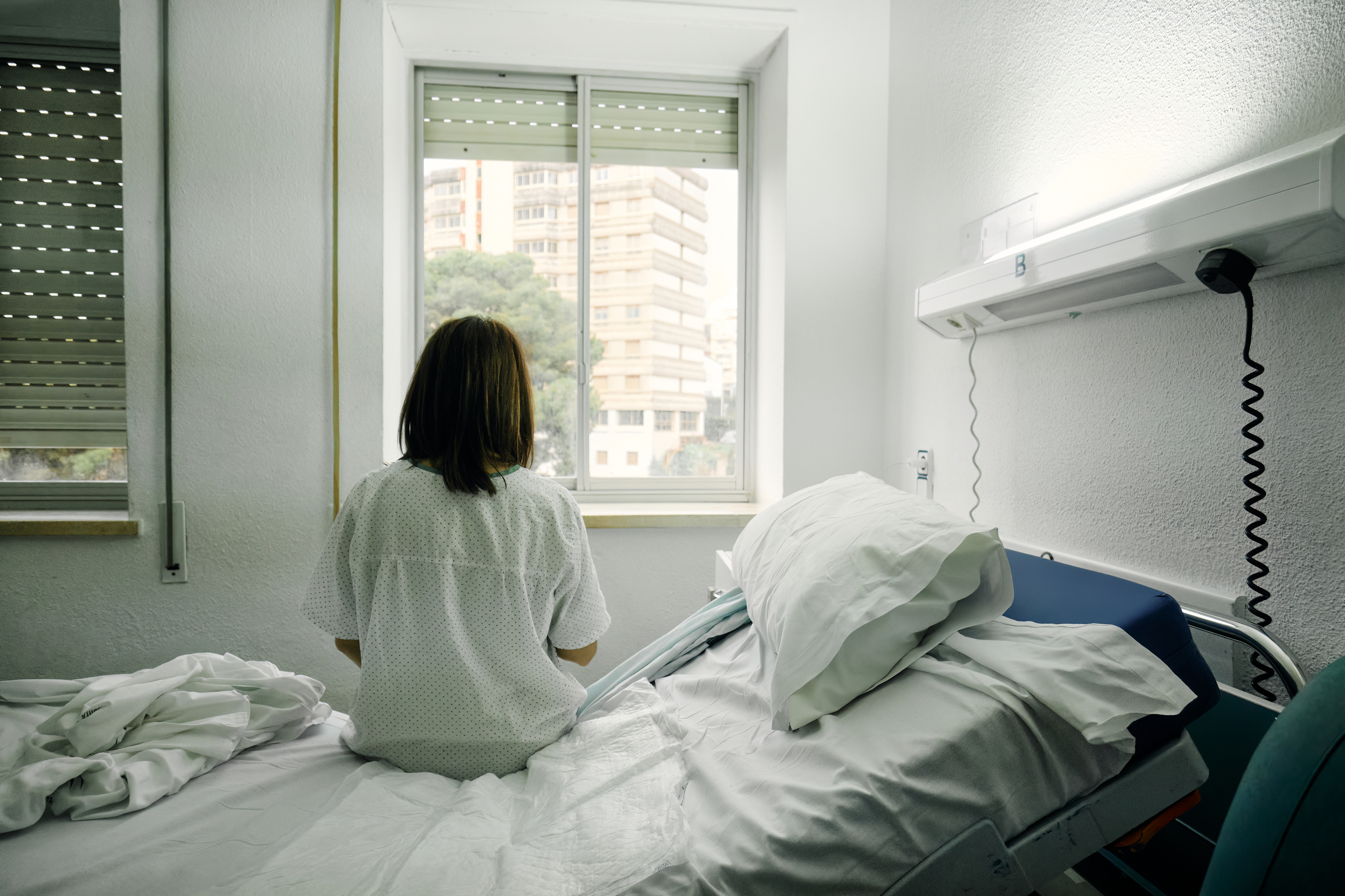 Person in a hospital gown sitting on a bed looking out of a window