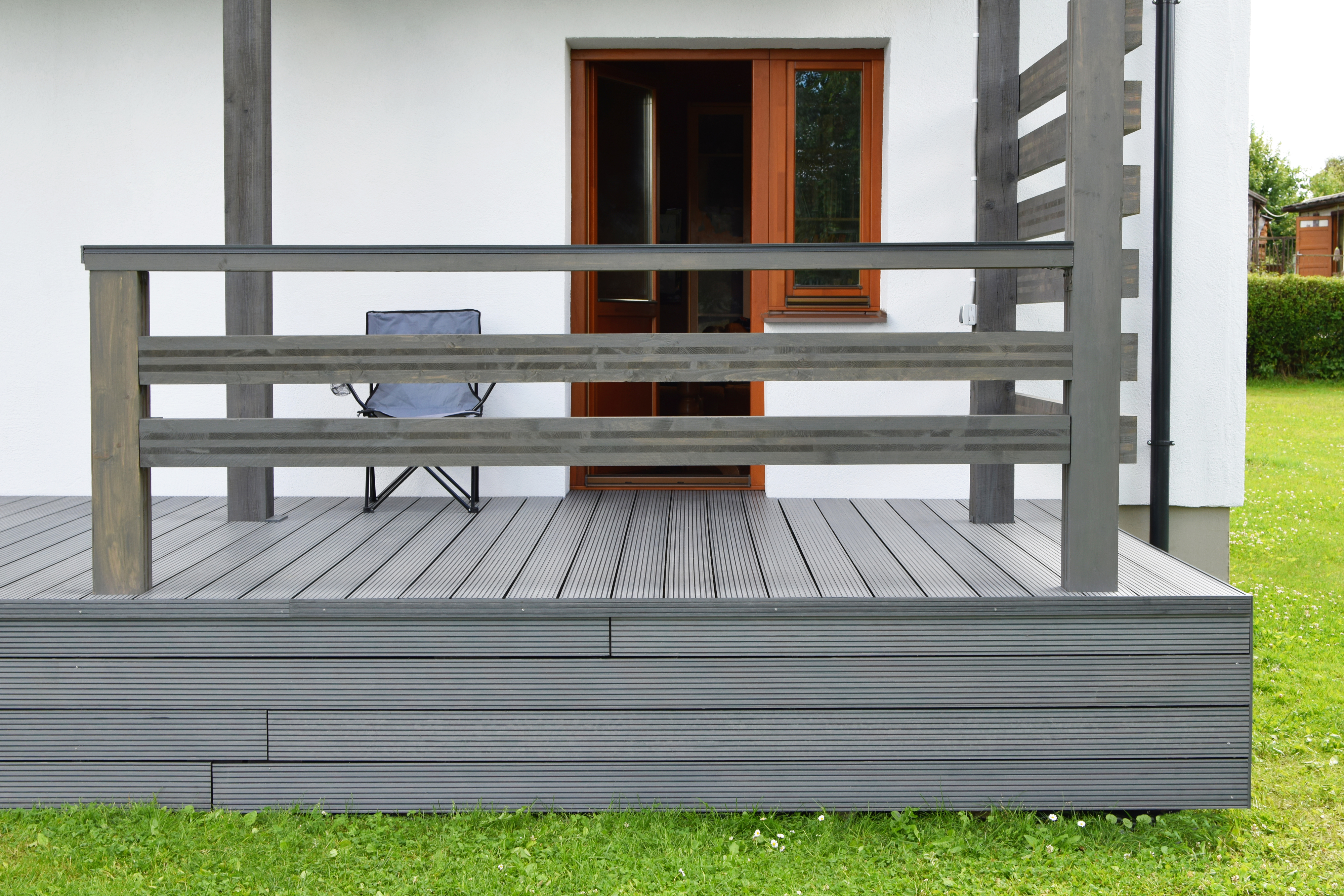 Wooden deck with modern railing in front of a house&#x27;s open door, including an outdoor chair on the deck