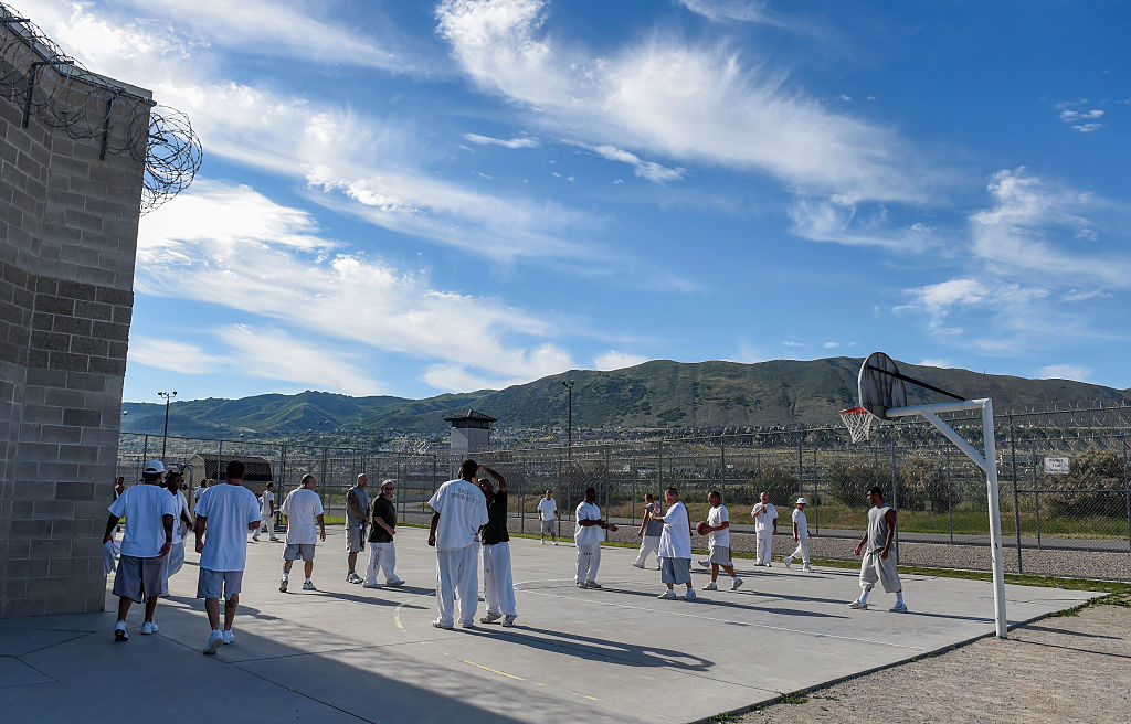 Inmates play basketball in a prison yard with guard towers in the background
