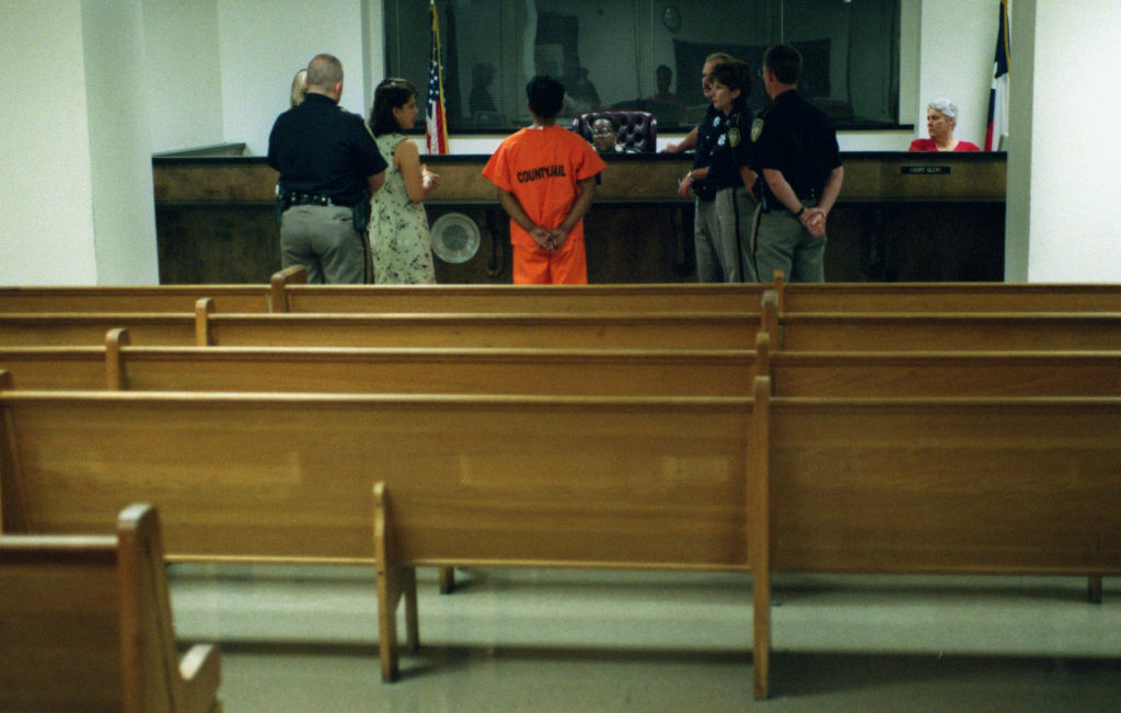 Ángel in an orange jumpsuit speaking with officers in a courtroom setting, with pews in the foreground