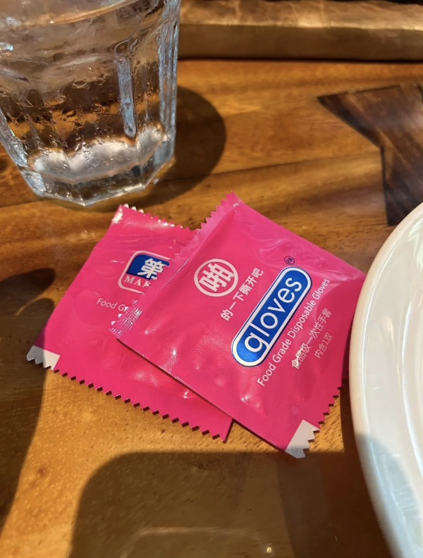 A packet of Aloves chili sauce on a wooden table beside a glass of water and edge of a white plate