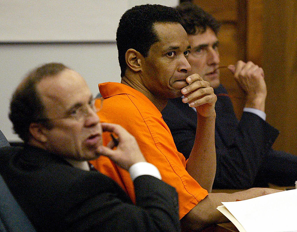 John Allen Muhammad in an orange jumpsuit with two others in suits, presumably his legal representation, in a courtroom