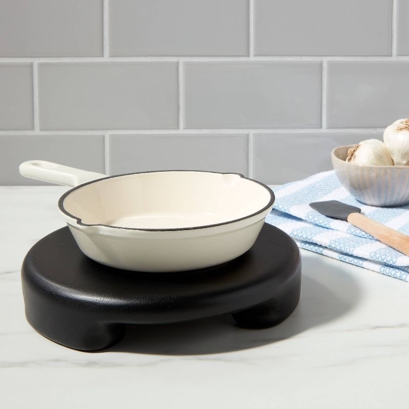 white enamel saucepan on a black trivet, beside garlic bulbs and kitchen utensils, on a kitchen counter