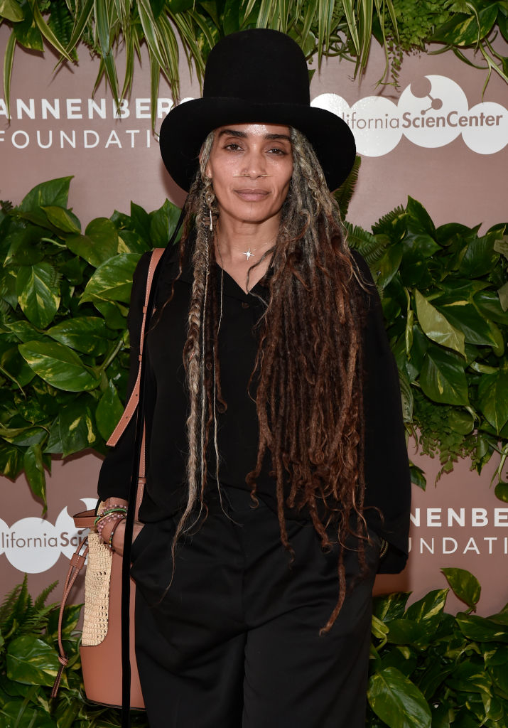 Lisa Bonet in a black outfit and hat, posing in front of a foliage backdrop