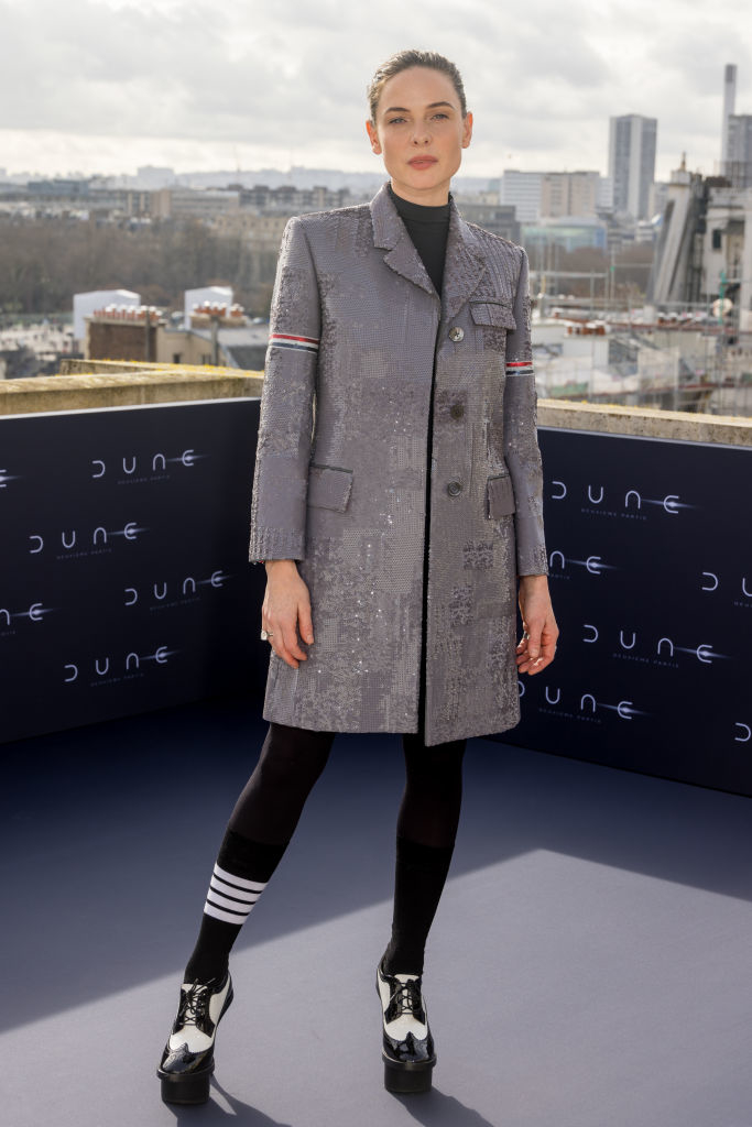 Rebecca in a textured coat and platform shoes standing before an event backdrop