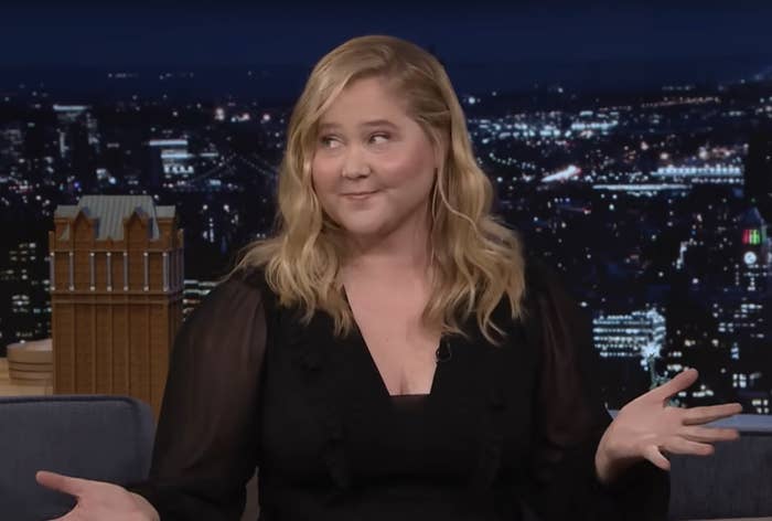 Amy Schumer in a talk show setting, gesturing with hands, dressed in a black outfit