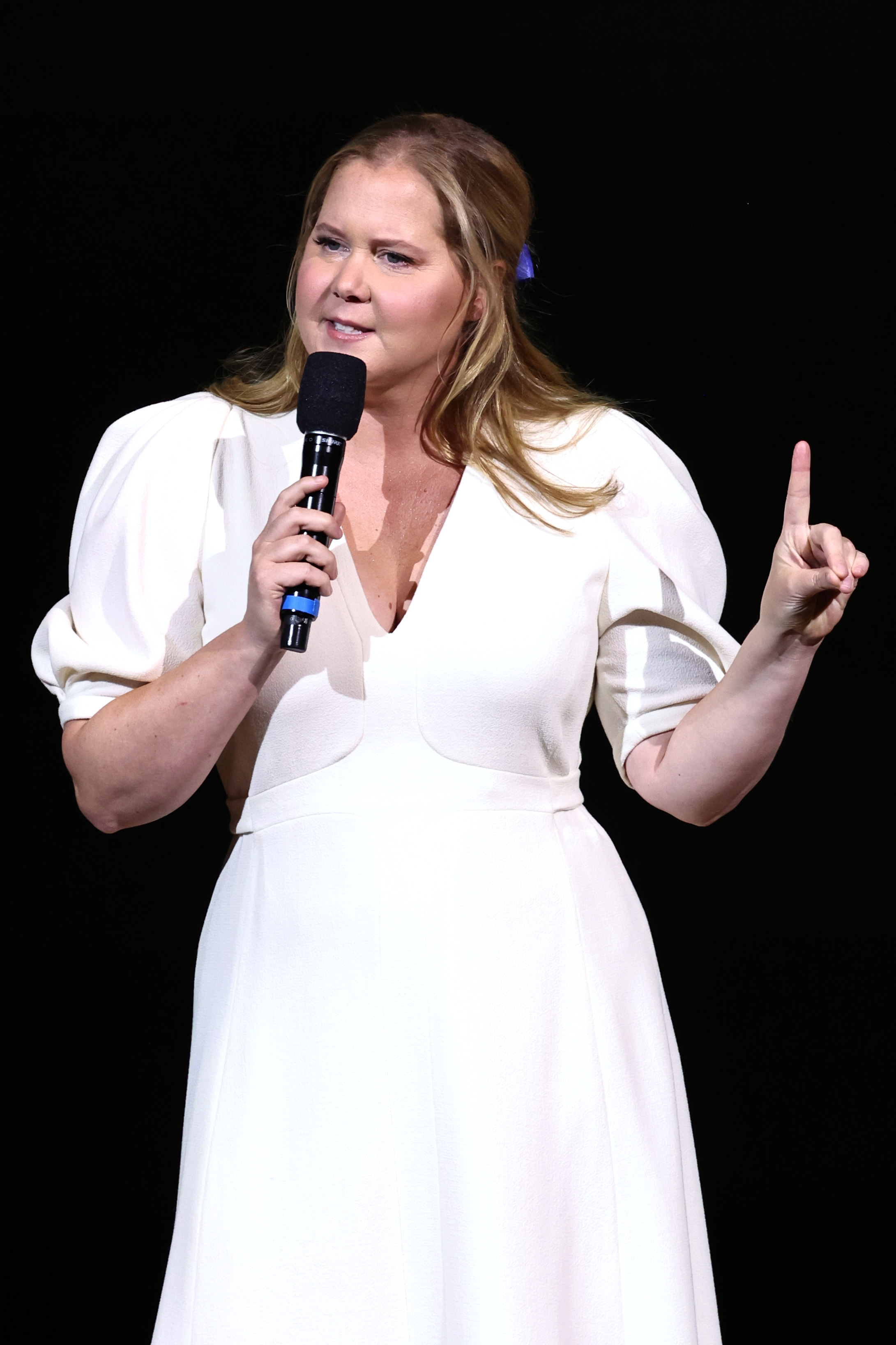 Amy, in a white dress with puffed sleeves, speaking into a microphone onstage