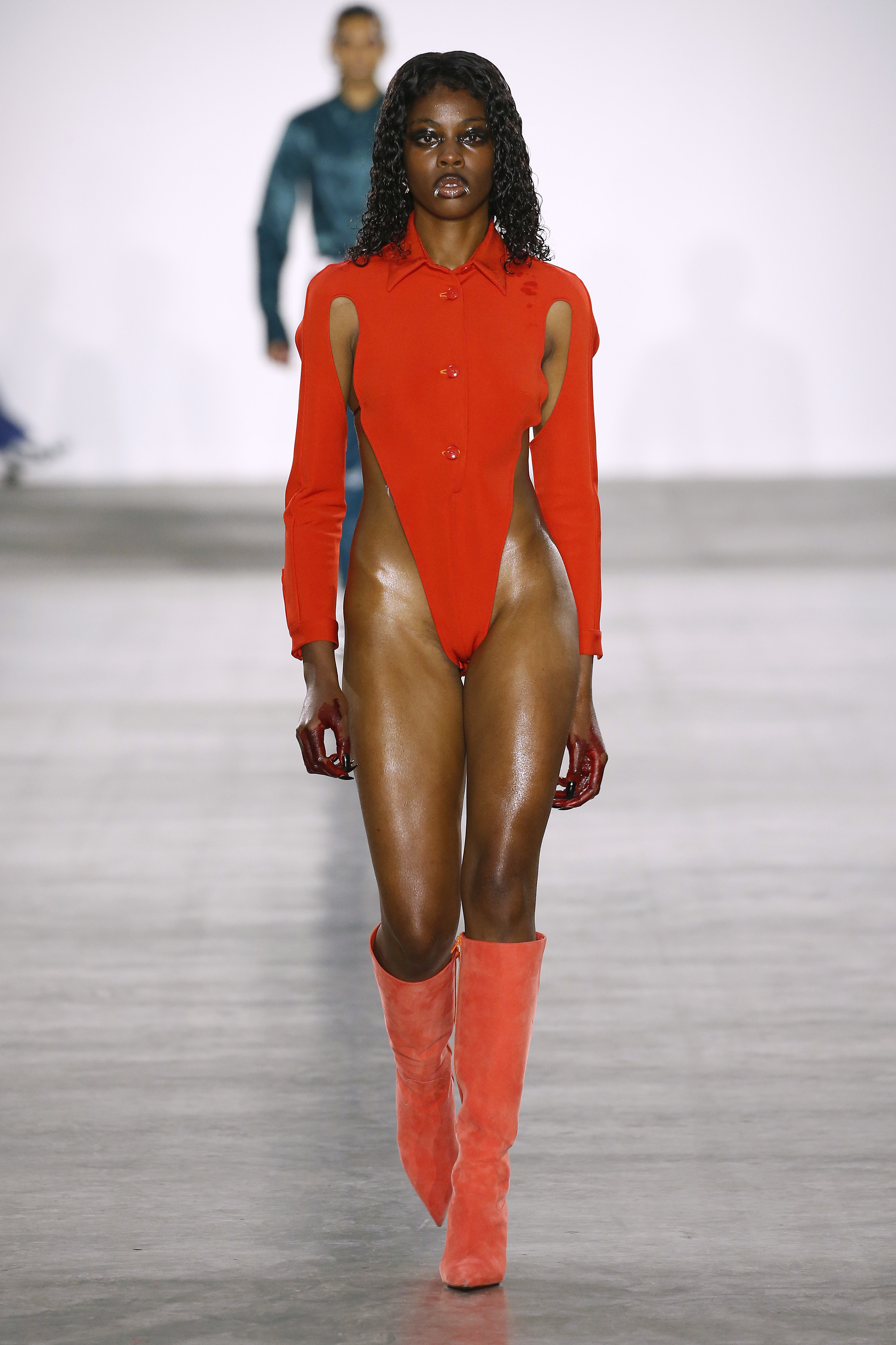 A model walks the runway in a high-cut red bodysuit and matching boots, another model in the background