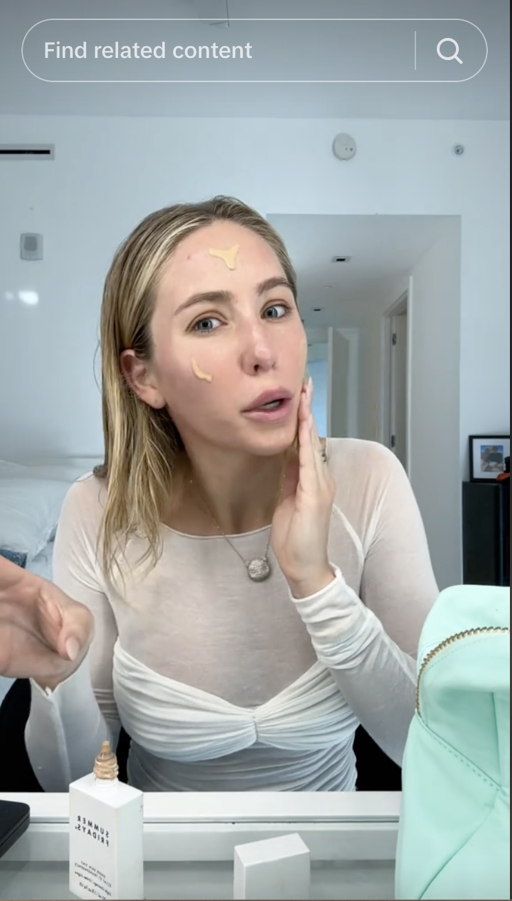 Woman applying skincare product in front of a mirror, with dots of the product on her face