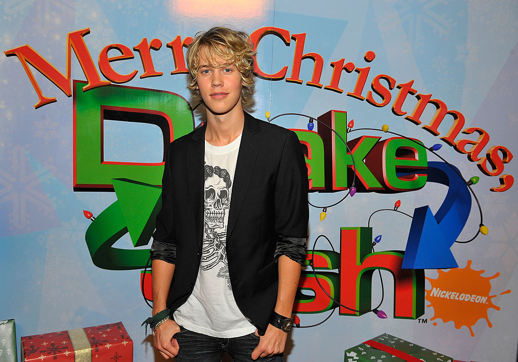 Austin with longish hair, standing before a &quot;Merry Christmas, Drake &amp;amp; Josh&quot; backdrop, wearing casual graphic tee and necklace