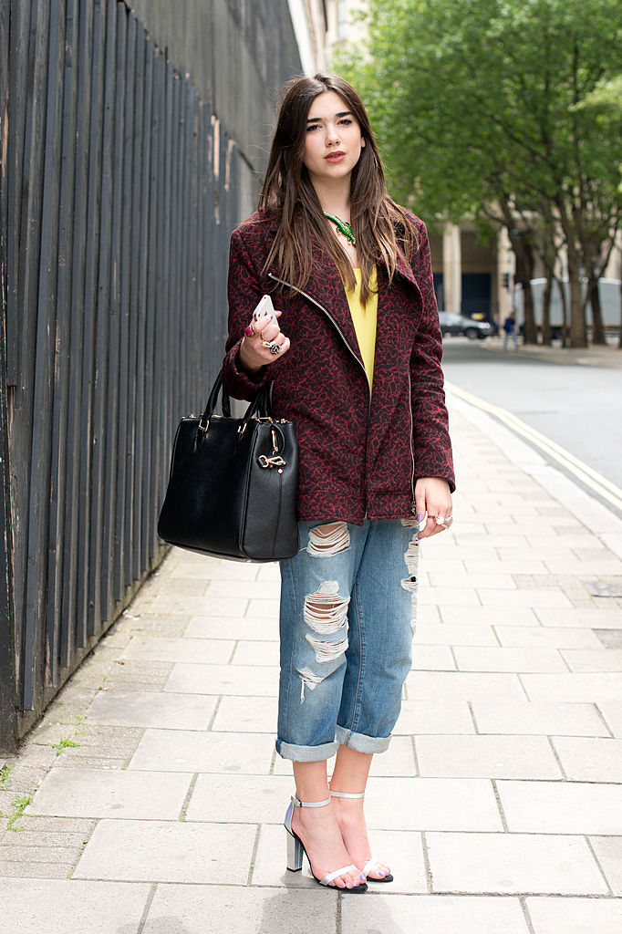 Dua in torn jeans and maroon jacket holding a black bag, standing on a sidewalk