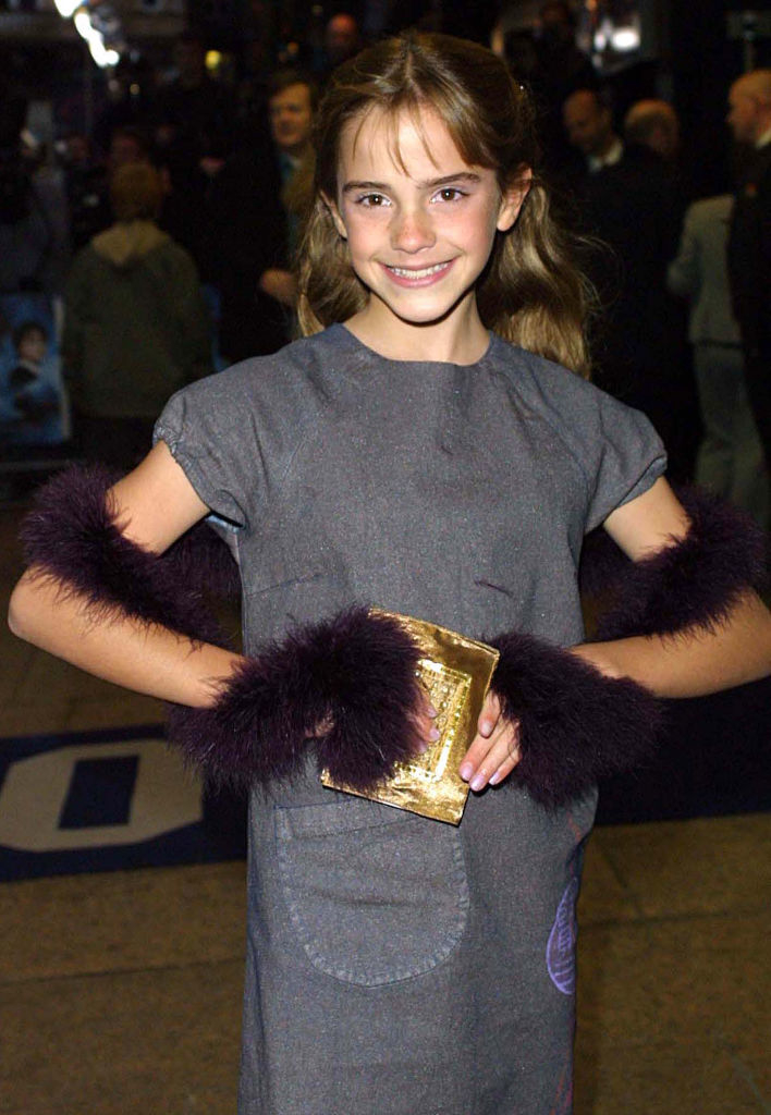 A young Emma in a gray dress with faux-fur cuffs, holding a gold clutch, at a movie premiere