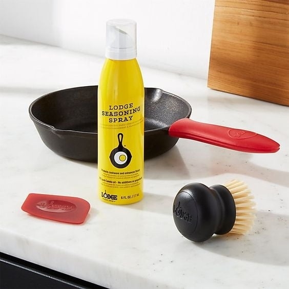 Seasoning spray bottle with cap, silicone brush, and pan scrapers on a countertop near a cast-iron skillet