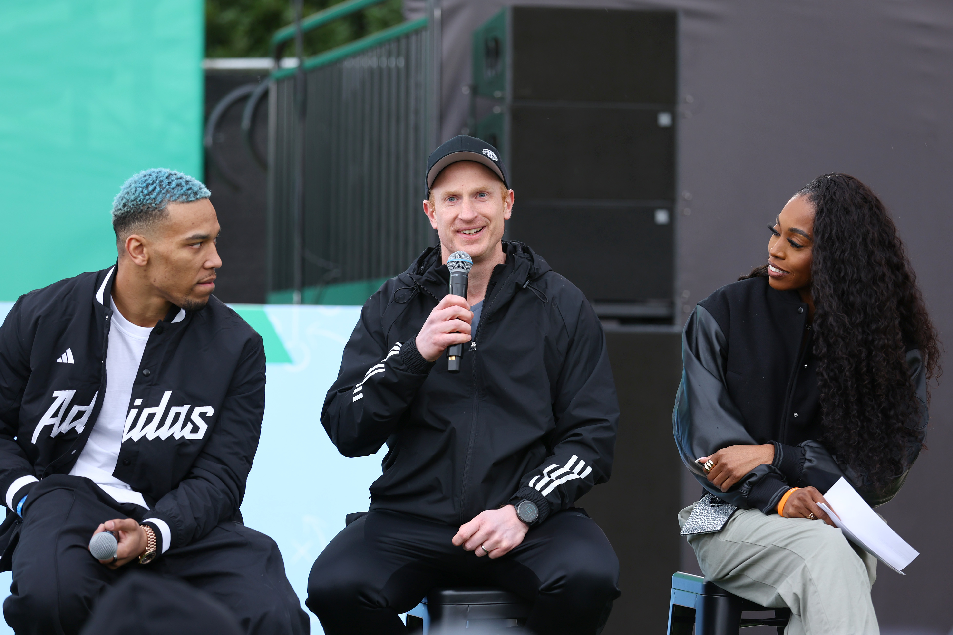 Three people in sportswear sitting and talking, one holding a microphone; outdoors, possibly at a sports event
