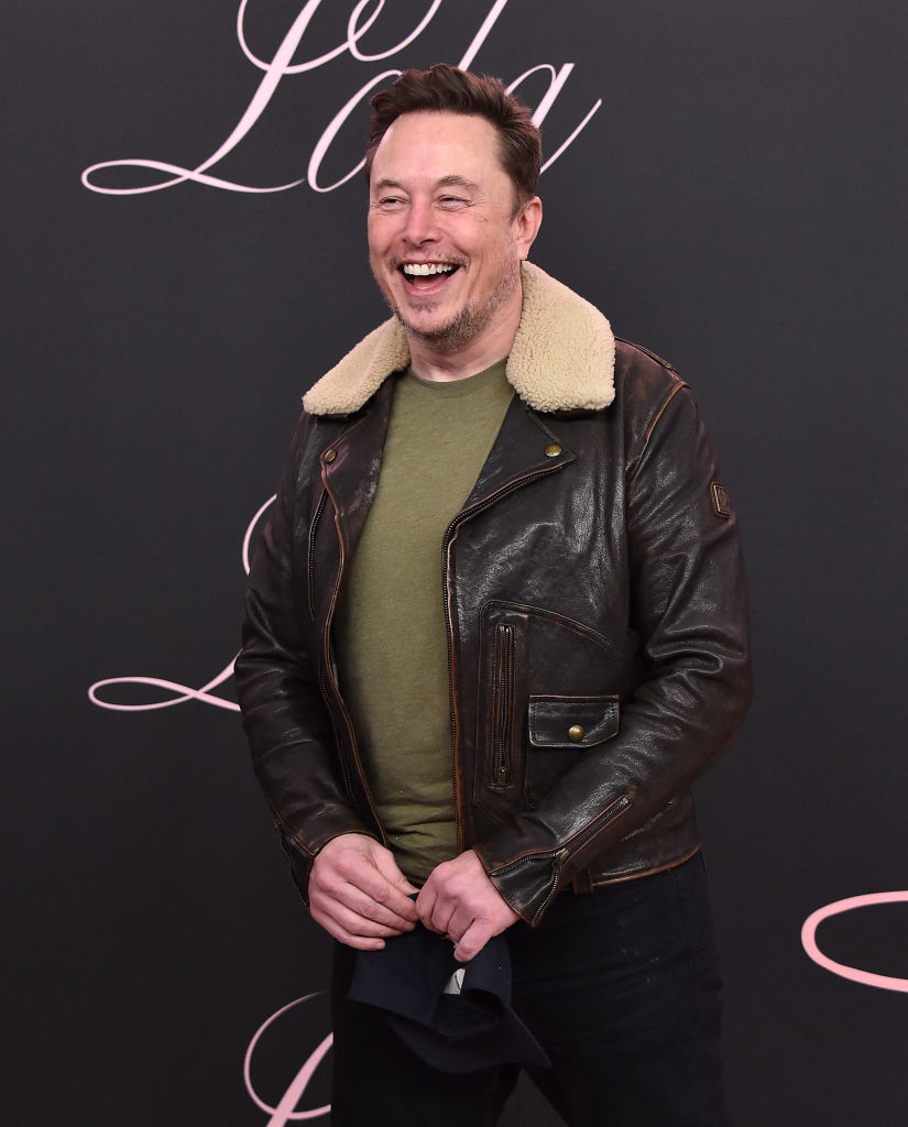 Elon laughing in a leather jacket with a shearling collar and a green T-shirt