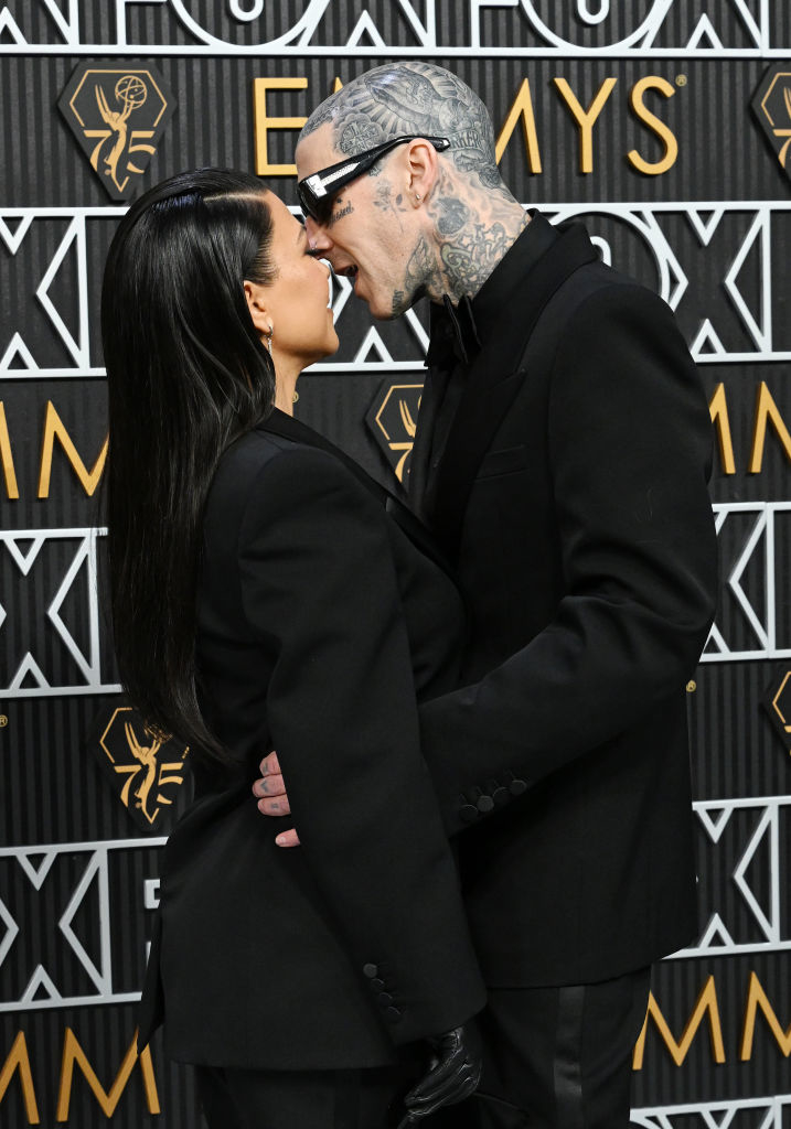 Kourtney Kardashian and Travis Barker share a kiss, both in black suits, at an event with patterned backdrop