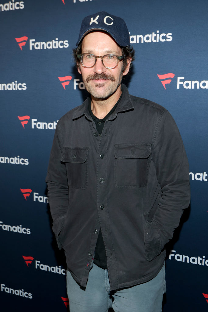 Paul with a mustache, in a cap and glasses, a loose shirt, and jeans, poses with hands in pockets at the Fanatics event