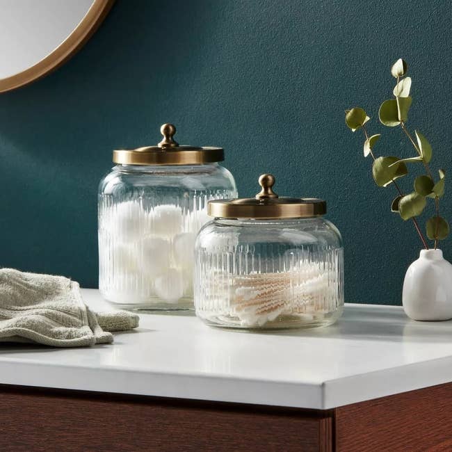 Two glass jars with golden lids on a table, one larger than the other, used for bathroom storage